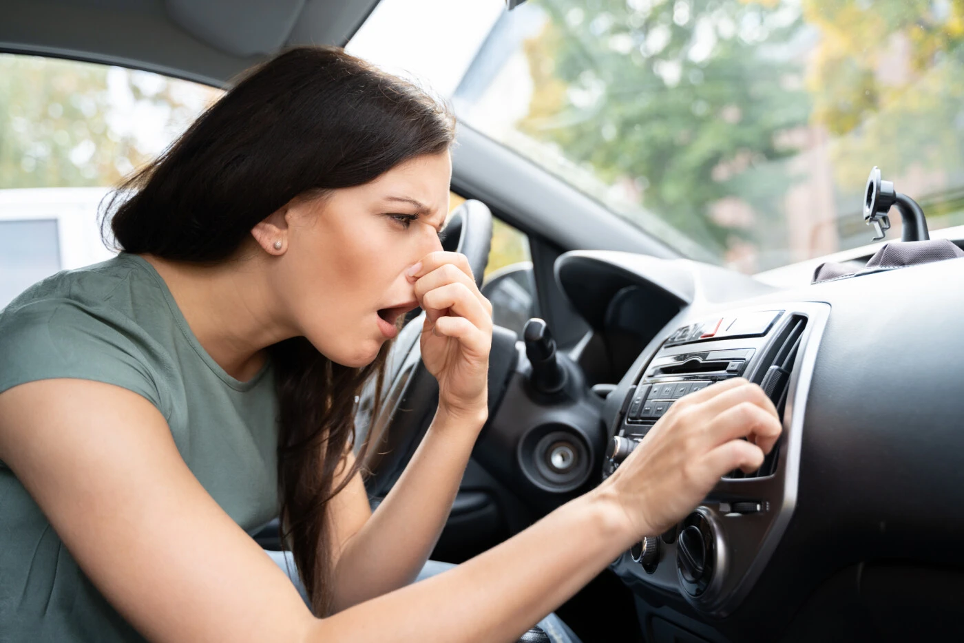 A woman drives while holding her nose, indicating a strong odor, possibly related to oil smelling like gas.