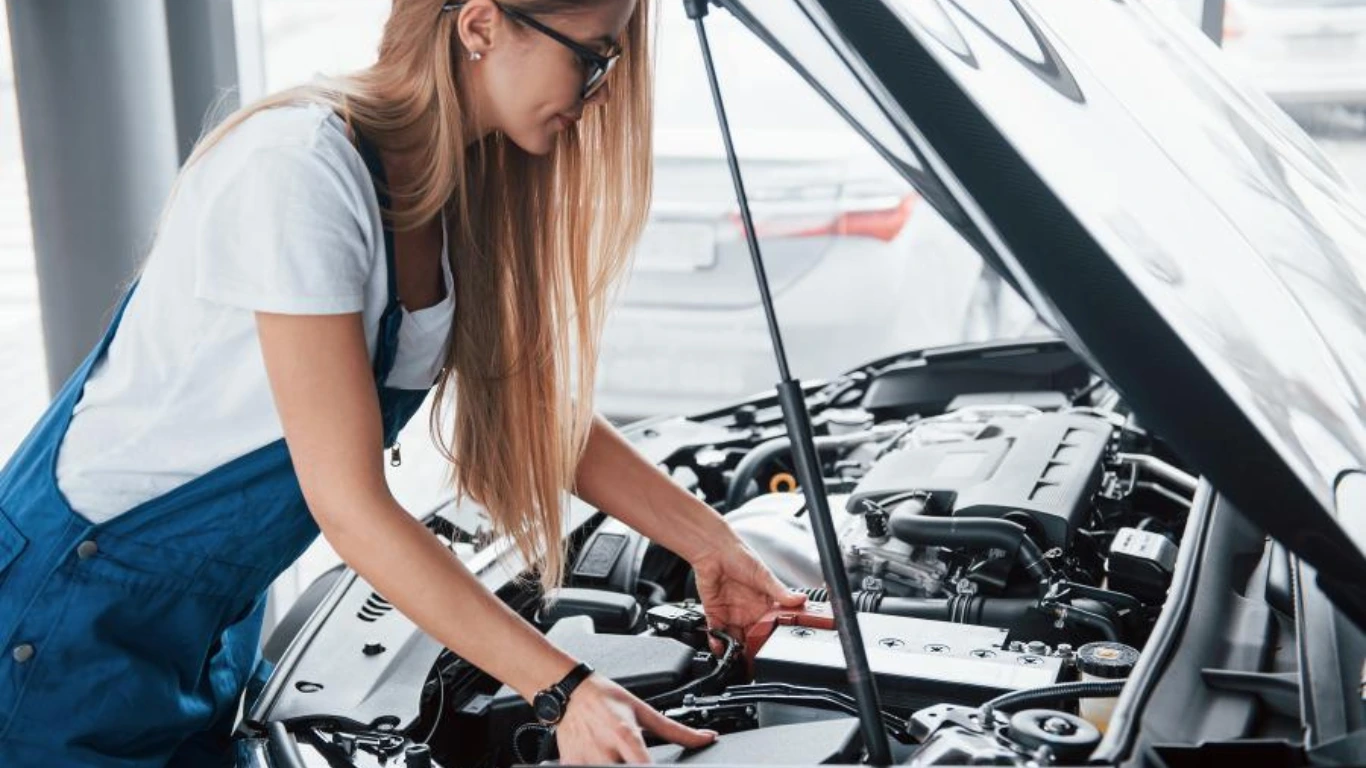 A woman in overalls diligently repairs a car engine, showcasing her mechanical skills and dedication to automotive work.